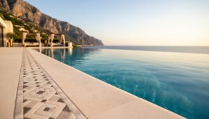 Wide view of a luxury infinity-edge swimming pool with mosaic tile details reflecting golden sunlight, blending into a Mediterranean horizon with sunlit cliffs in the background.